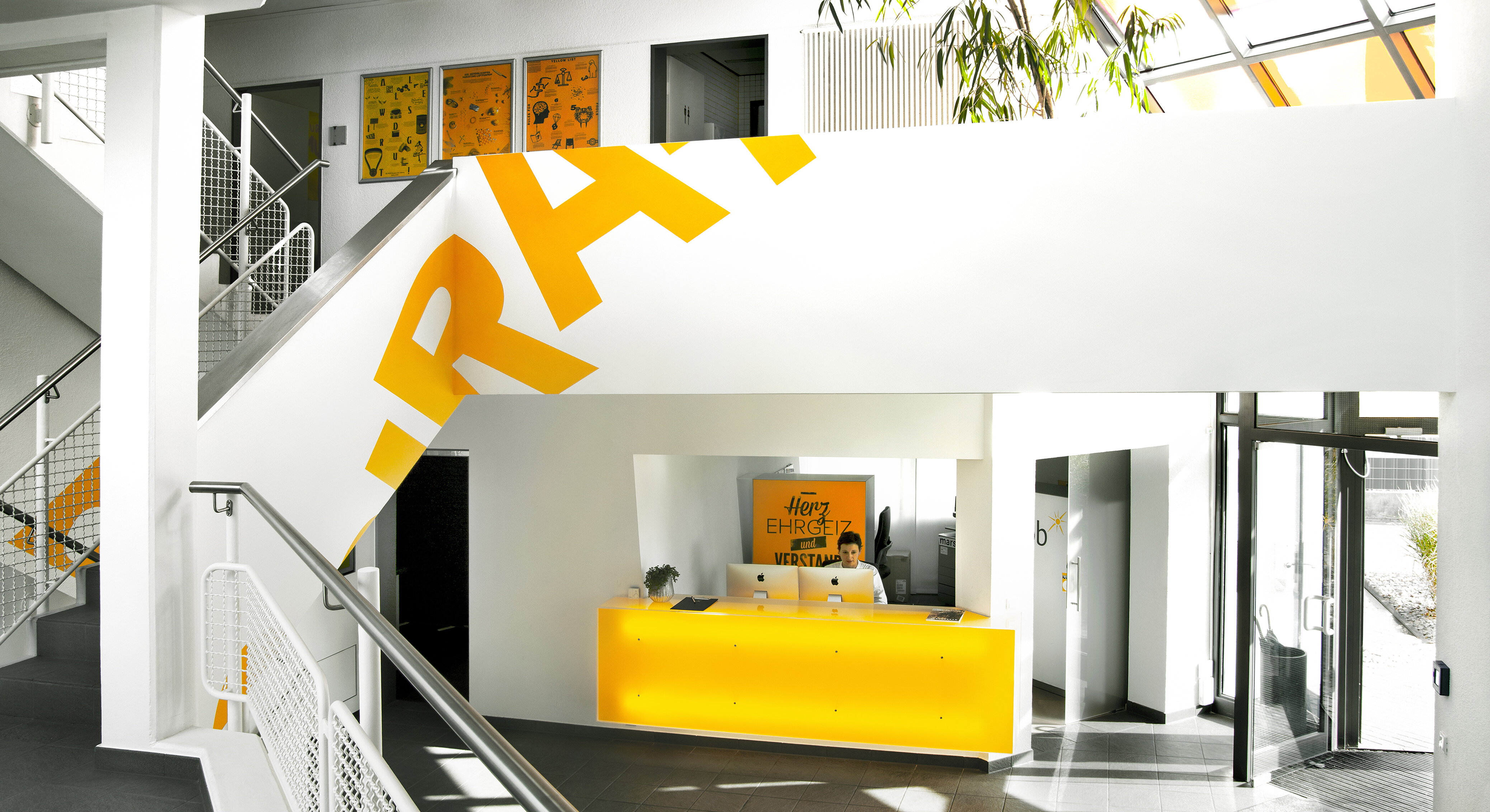 The picture shows the bright reception hall of wob AG from the inside with a view of the reception desk. A yellow reception desk can be seen in the centre, behind which a person is sitting at a computer. On the far right of the picture, the building entrance can be recognised as a glass door. On the left is an open staircase with parts of the word Inspiration in yellow letters on the side walls. | © wob AG