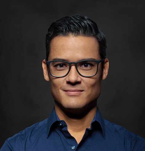 Dieses Bild ist ein Porträt von Dennis Güth. Er trägt eine Brille und ein dunkel blaues Hemd. Seine Arme sind verschränkt. Der Hintergrund ist dunkel. | © wob AG